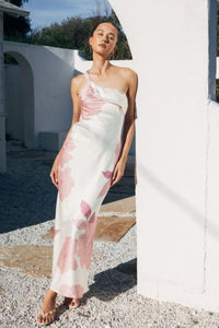Woman in a one-shoulder floral dress standing outdoors near a white wall.