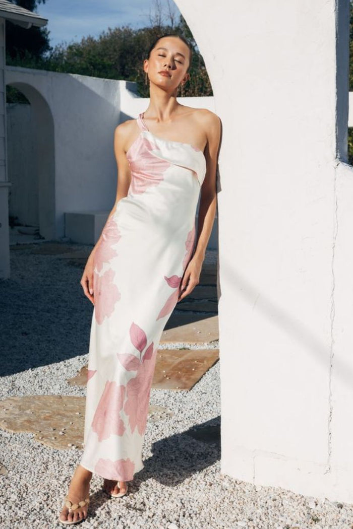 Woman in a one-shoulder floral dress standing outdoors near a white wall.