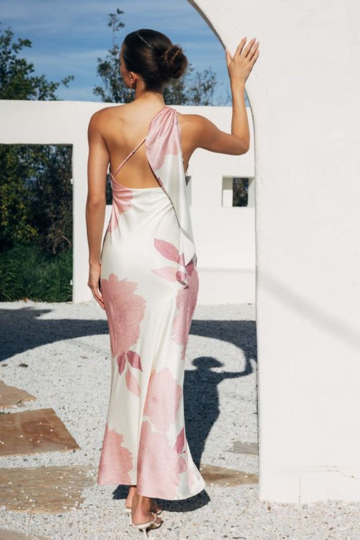 Woman wearing a floral one-shoulder dress standing against a white wall with greenery.