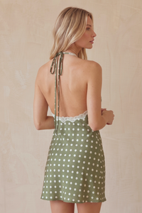 A woman shown from the back wearing a sage green halter mini dress with white polka dots and a lace back trim, set against a neutral beige background.