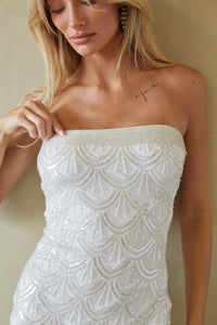 A close-up of a woman wearing a strapless white beaded dress with a scalloped, sequined pattern against a neutral beige background.