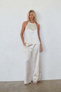 Woman wearing a white sleeveless top and wide-leg pants against a plain white wall.