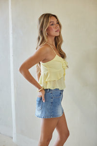 Woman wearing a yellow ruffled top and denim skirt against a white wall