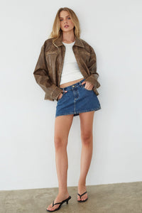 Woman wearing a brown leather jacket, white top, and blue denim skirt against a white background