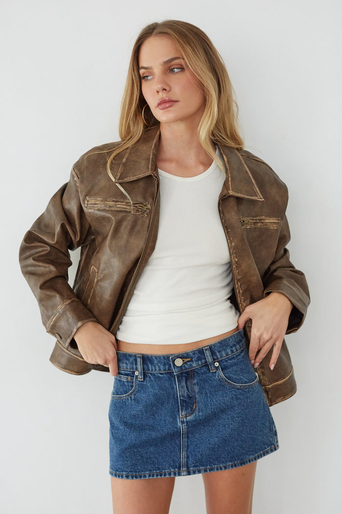 Woman wearing a brown leather jacket, white top, and blue denim skirt against a white background