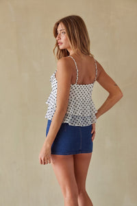 A woman modeling a white ruffled camisole top with black polka dots, paired with a blue denim mini skirt, photographed against a neutral beige textured wall.