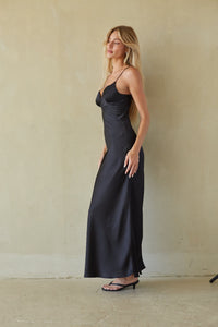 A woman modeling a black satin maxi dress with spaghetti straps and a sweetheart neckline, standing against a textured beige plaster wall.