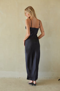 A woman wearing a black satin maxi slip dress with thin spaghetti straps, shown from the back against a textured beige plaster wall.