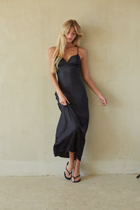 A woman with long blonde hair models a black satin slip dress with thin spaghetti straps and a draped, gathered skirt, standing against a textured beige plaster wall.