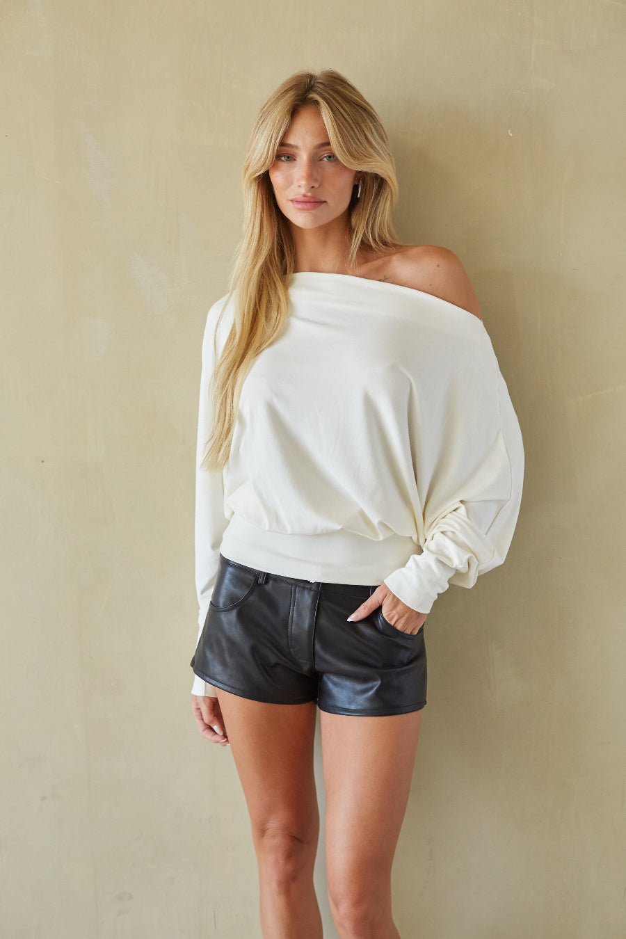 A woman modeling an off-the-shoulder cream long-sleeve top and black faux-leather shorts against a textured beige wall.