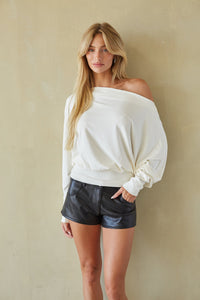 A woman modeling an off-the-shoulder cream long-sleeve top and black faux-leather shorts against a textured beige wall.