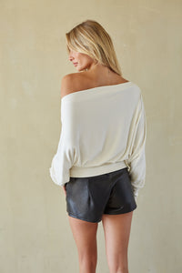 A woman shown from the back wearing an off-the-shoulder cream long-sleeve top and black faux-leather shorts, standing against a textured beige wall.