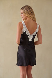 A woman shown from the back wearing a black satin slip dress with white lace trim along the open back and shoulder straps, standing against a plain beige wall.