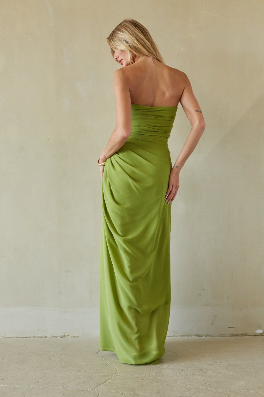 A woman wearing a strapless olive green maxi dress, photographed from behind against a neutral beige plaster wall and floor.