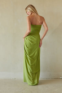 A woman wearing a strapless olive green maxi dress, photographed from behind against a neutral beige plaster wall and floor.