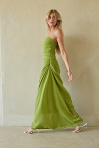 A woman modeling a strapless olive green maxi dress with ruched detailing and a flowing skirt, paired with clear high-heeled sandals, standing against a textured beige wall.