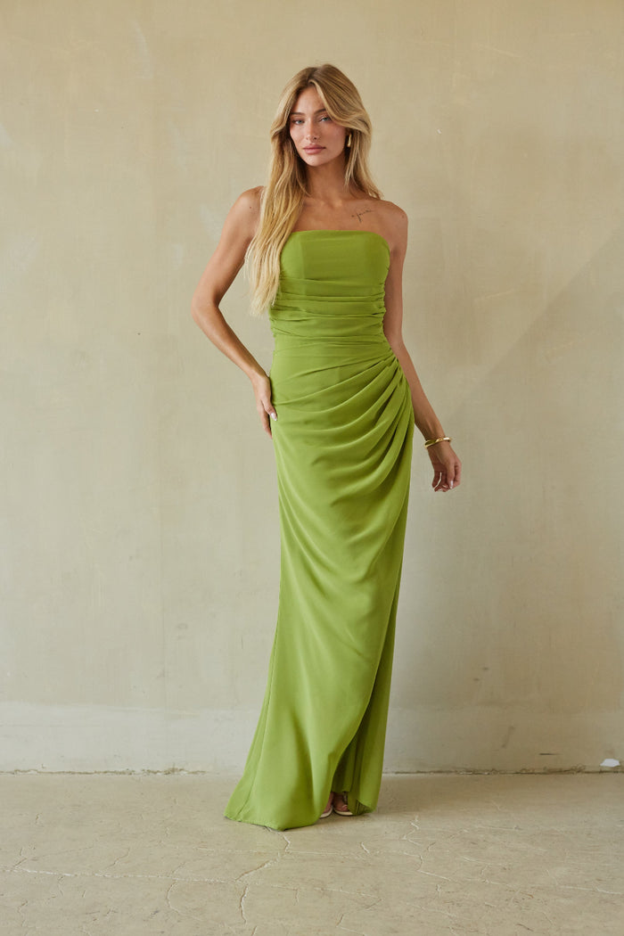 A woman modeling a strapless lime green maxi dress with draped detailing, standing against a textured beige plaster wall.