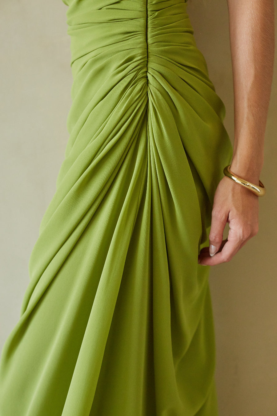 Close-up of a draped olive green dress with a gathered waist and a model's hand wearing a gold bangle, set against a neutral beige background.