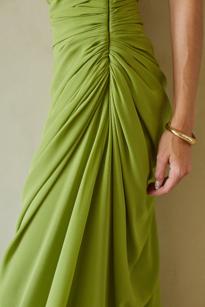 Close-up of a draped olive green dress with a gathered waist and a model's hand wearing a gold bangle, set against a neutral beige background.