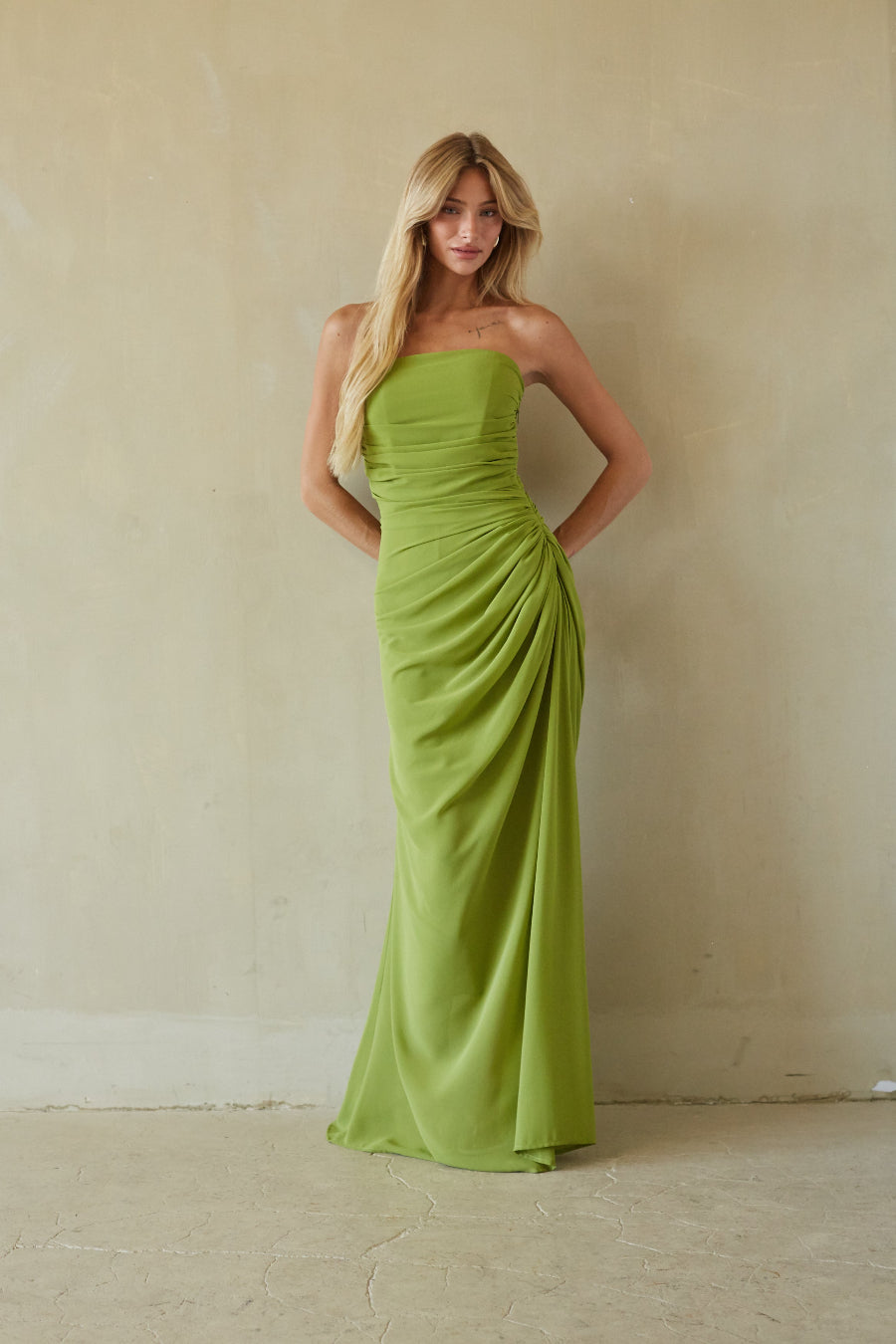 A woman modeling a strapless lime green floor-length gown with gathered draping, standing against a textured beige plaster wall.