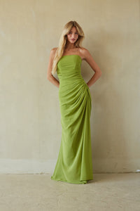 A woman modeling a strapless lime green floor-length gown with gathered draping, standing against a textured beige plaster wall.