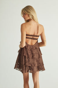 A woman shown from the back wearing a brown strapless mini dress with a ruffled tiered skirt and double horizontal cutout straps across the upper back, against a plain light gray studio background.