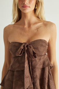A close-up of a model wearing a strapless brown ruffled top with a tonal floral jacquard pattern and a large bow at the bust.