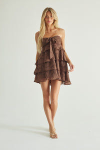 A blonde model wearing a brown strapless ruffled mini dress with a bow at the bust, standing against a plain light gray studio background.