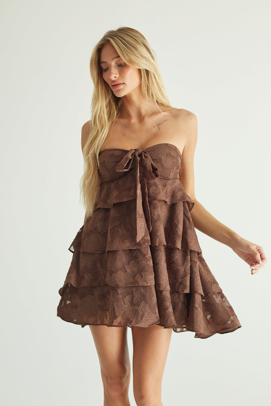A blonde model wearing a brown strapless tiered mini dress with a bow detail at the bust, photographed against a plain light gray studio background.