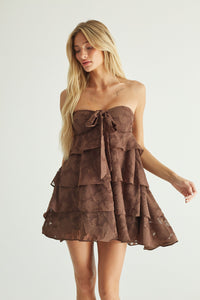 A blonde model wearing a brown strapless tiered mini dress with a bow detail at the bust, photographed against a plain light gray studio background.
