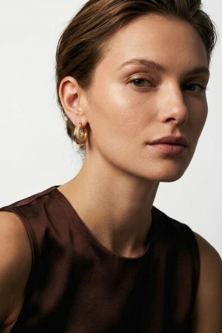 A close-up portrait of a woman wearing a small gold hoop earring, shown from the chest up against a clean light gray background.