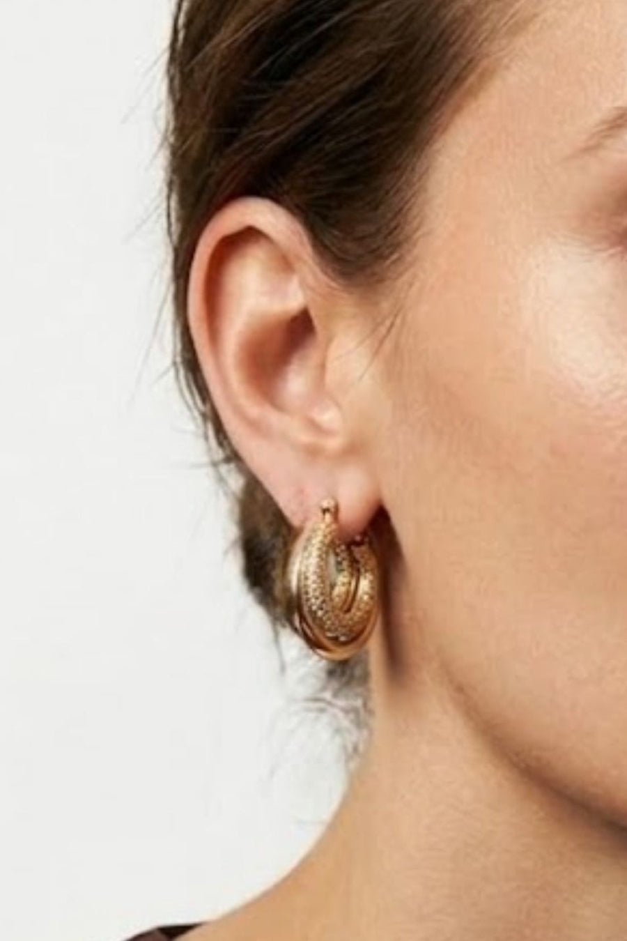 A close-up of a woman's ear wearing a textured gold hoop earring against a plain light background.