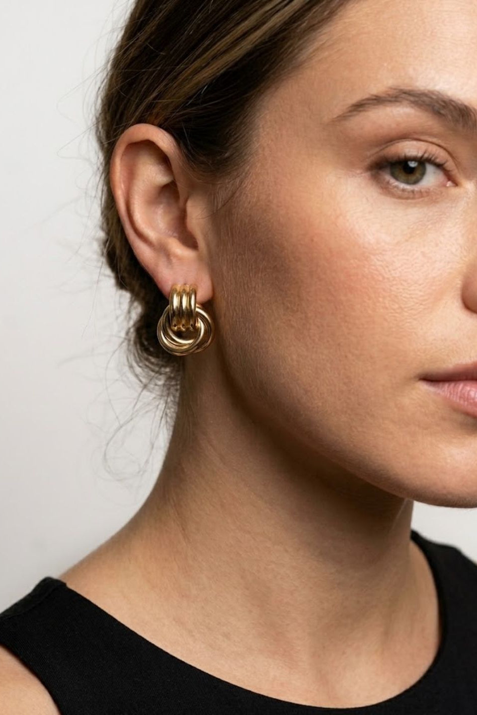 A close-up of a woman wearing a pair of gold triple-ring hoop earrings, shown from the side against a plain light gray background.
