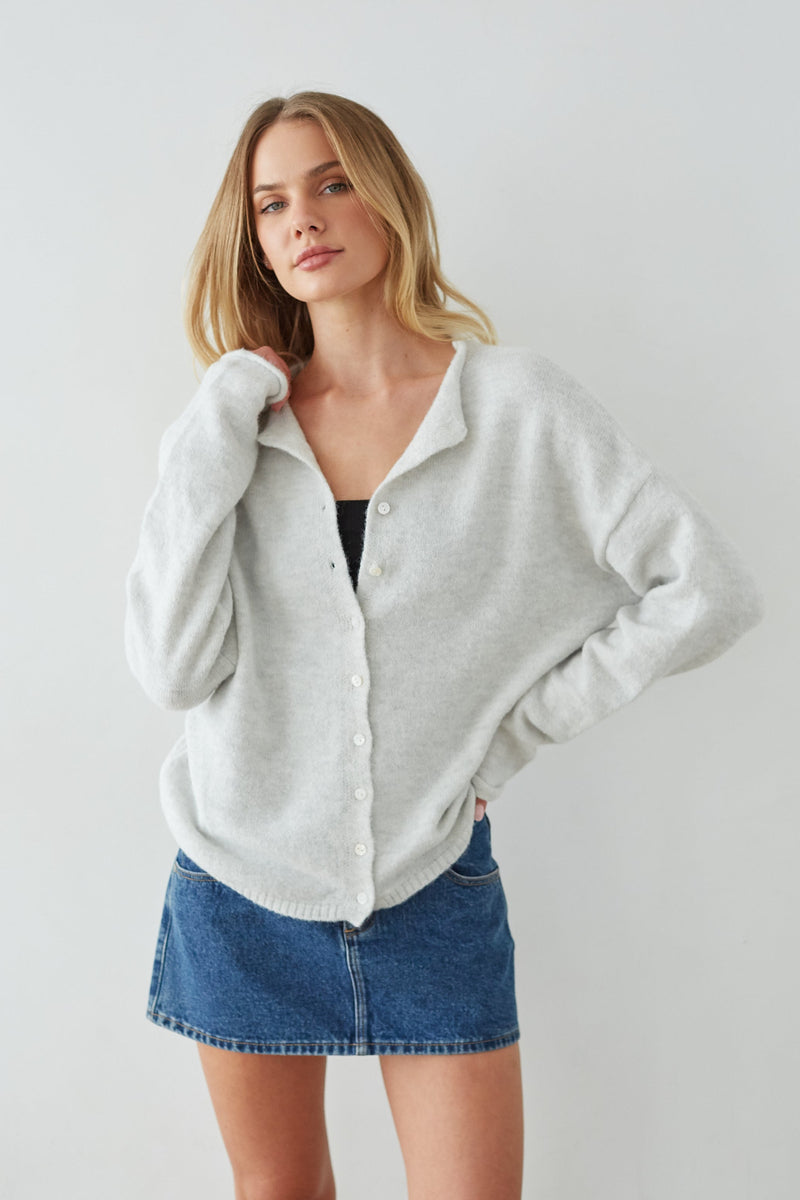 Woman wearing a light gray cardigan over a black top and blue denim skirt on a plain background