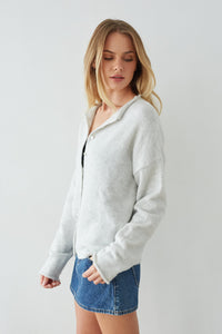 Woman wearing a light gray button-up sweater and denim shorts on a plain background