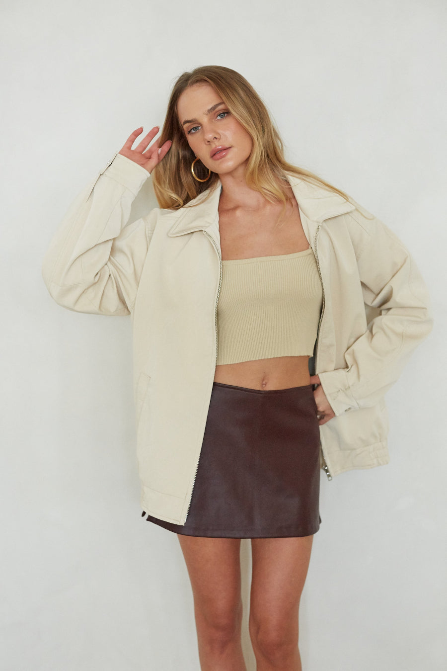 Woman wearing a beige jacket, beige top, and brown leather skirt against a white background
