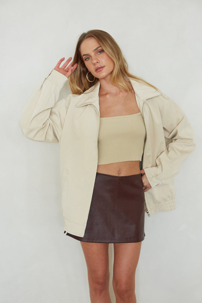 Woman wearing a beige jacket, beige top, and brown leather skirt against a white background