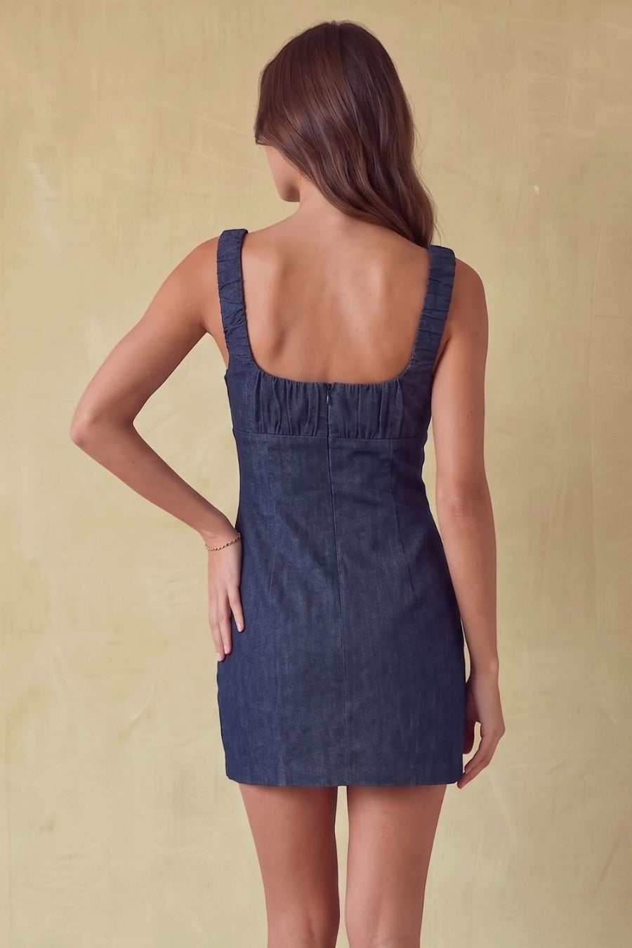 A woman shown from the back wearing a dark blue denim mini dress with a square neckline and shoulder straps, standing against a textured beige background.