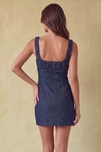 A woman shown from the back wearing a dark blue denim mini dress with a square neckline and shoulder straps, standing against a textured beige background.