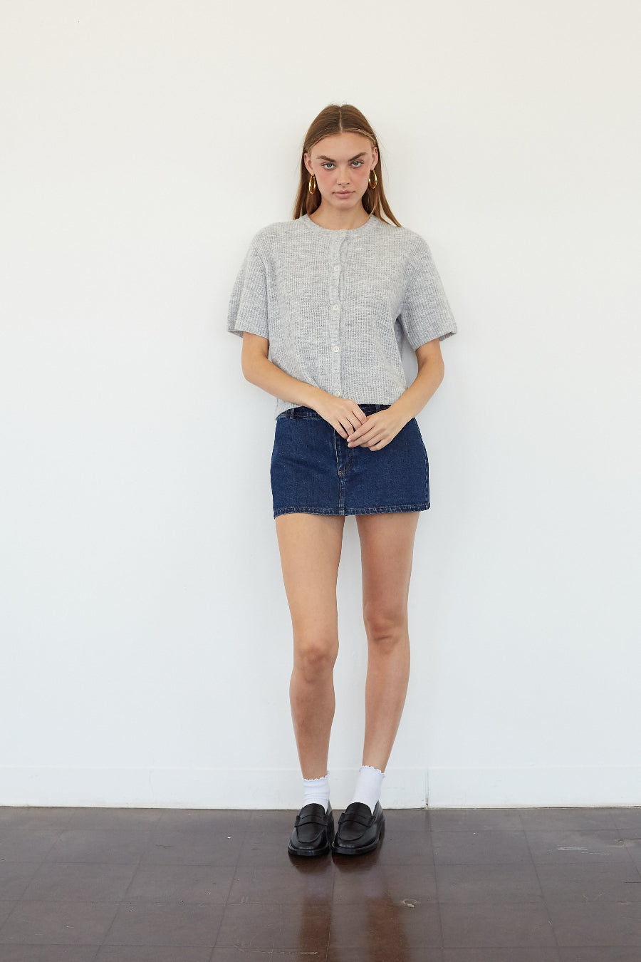 A woman stands against a white wall wearing a light gray short-sleeve knit top, a dark blue denim mini skirt, white socks, and black loafers.