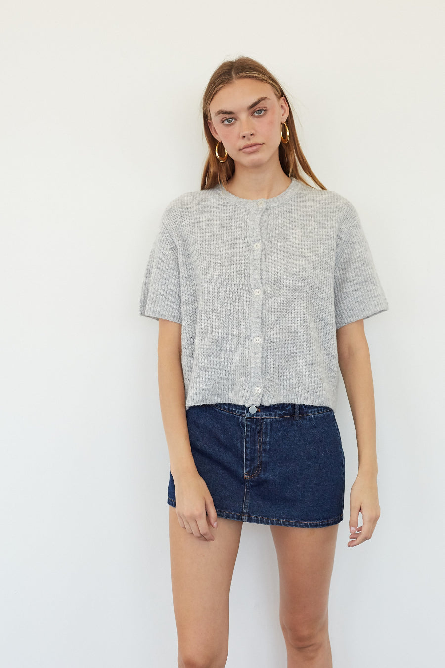 A woman modeling a light gray short-sleeve cropped button-front top with a textured knit pattern, paired with a dark denim mini skirt, against a plain white background.