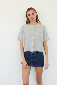 A woman modeling a light gray short-sleeve cropped button-front top with a textured knit pattern, paired with a dark denim mini skirt, against a plain white background.