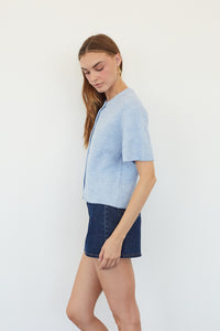 A woman modeling a light blue short-sleeve textured knit top with dark blue denim shorts against a plain white studio background.
