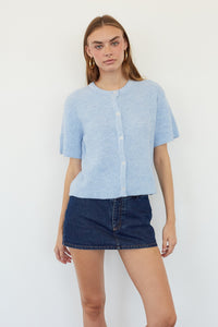 A woman modeling a light blue short-sleeve button-front knit top with a dark blue denim mini skirt, standing against a plain white background.