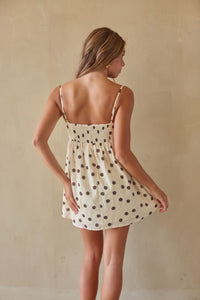 A woman wearing a short cream-colored spaghetti-strap dress with dark brown polka dots, shown from behind against a neutral beige wall.