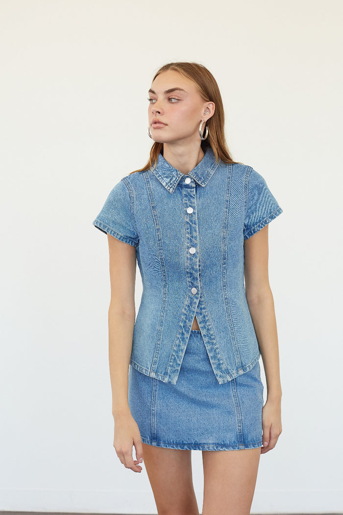 A woman modeling a light blue denim short-sleeve button-front shirt and matching denim mini skirt against a plain white studio background.