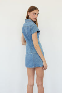 A woman modeling a light blue denim short-sleeve mini dress from the back, standing against a plain white studio background.