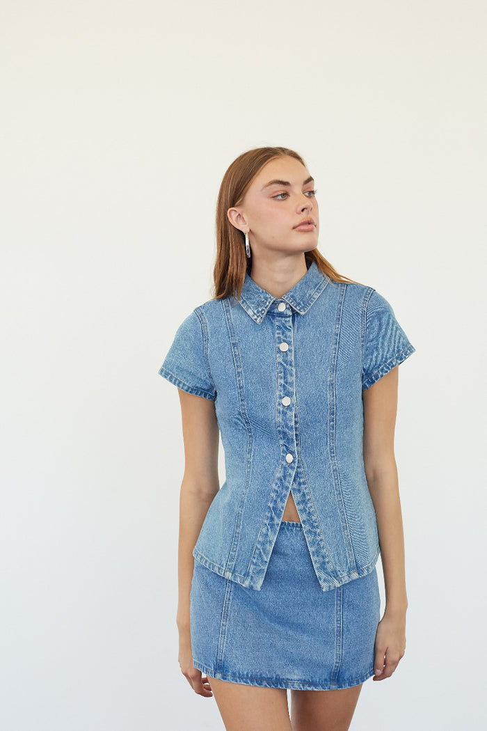 A woman modeling a light blue denim short-sleeve button-up shirt and matching denim mini skirt against a plain white studio background.