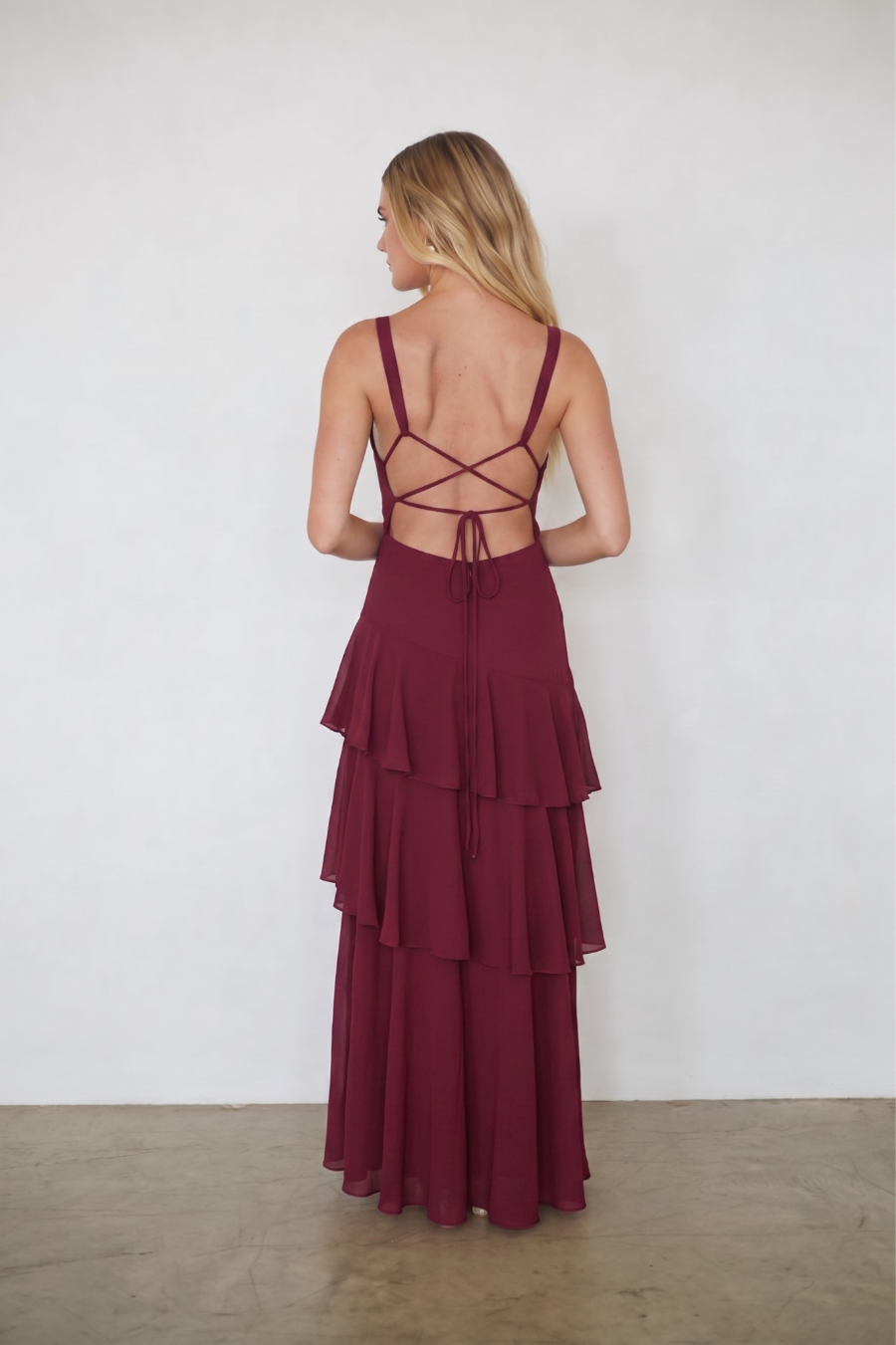 A woman stands with her back to the camera wearing a long burgundy maxi dress with a strappy open back and tiered ruffle skirt, photographed against a plain white wall and concrete floor.
