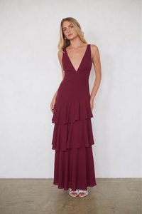 A woman modeling a sleeveless, deep V-neck maxi dress in burgundy with a tiered ruffle skirt, standing against a plain white wall.